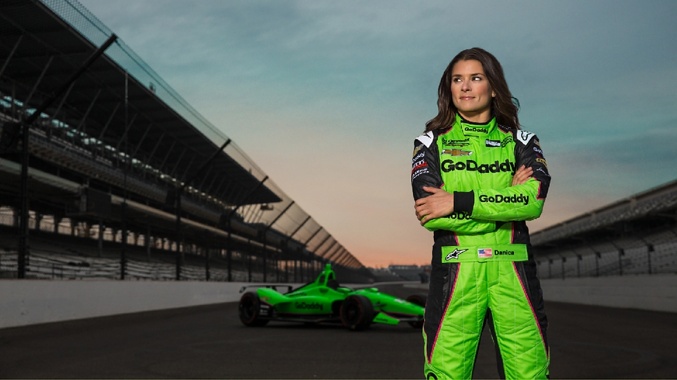 A woman in a bright green racing suit stands confidently on a racetrack at sunset. A green race car is visible in the background.