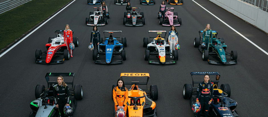 Race cars lined up on a track with drivers standing beside them. Vibrant colors and sponsor logos visible. Stadium seating in the background.