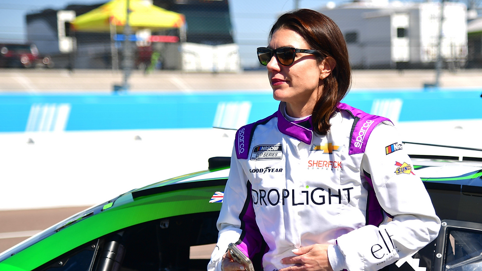 A woman in a racing suit with "DROPLIGHT" text stands beside a green car on a race track. She's wearing sunglasses and appears focused.