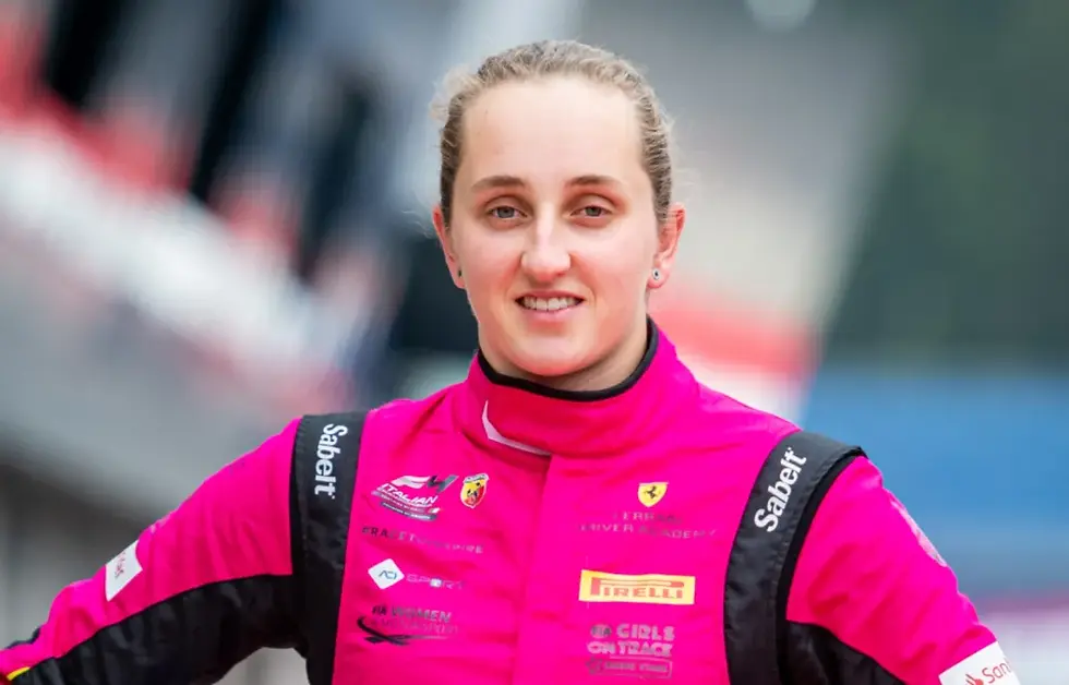 Smiling woman in a bright pink Ferrari racing suit with sponsor logos, standing at a racetrack with a blurred background.