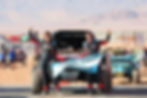 Two people in racing suits celebrate with a "General Cabrera Cordoba" flag in front of a red race car. Desert and other cars in background.