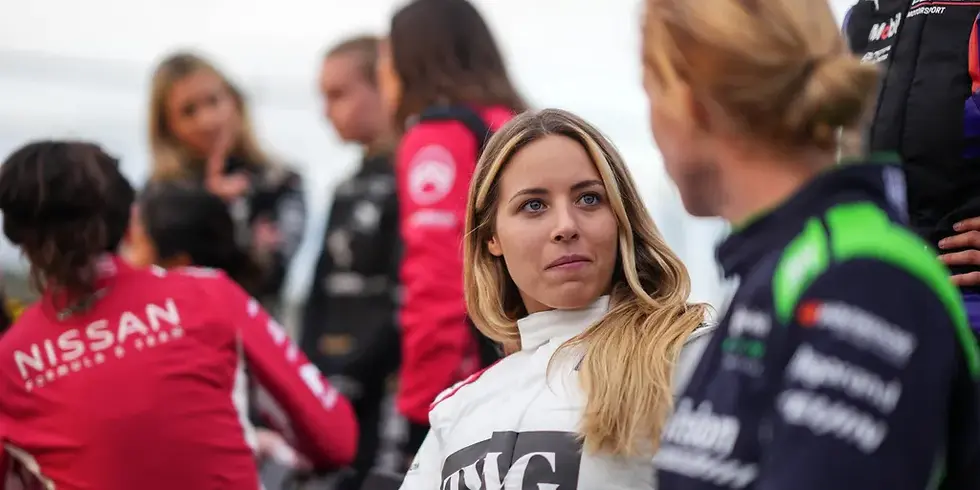 Women in racing suits talk on a motorsport grid, with a blonde woman in white listening; Nissan logo visible.