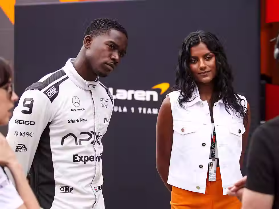 A man in a race suit stood next to a  woman. They are in the F1 paddock. 