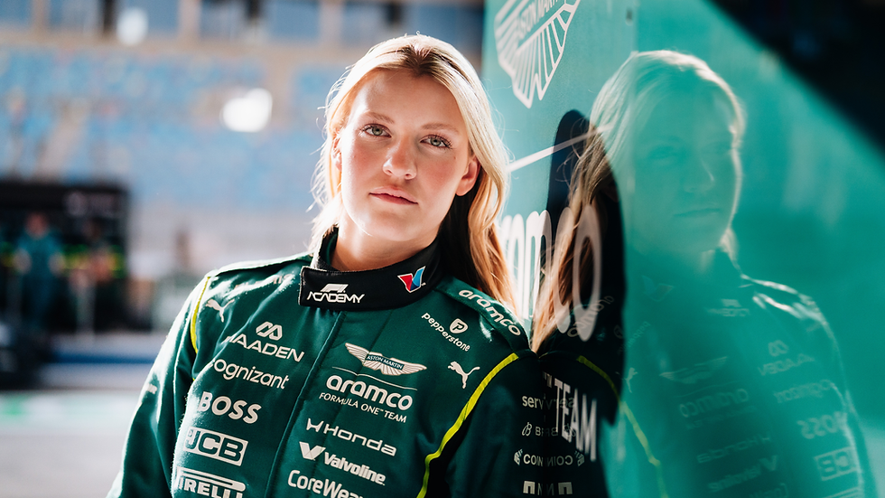Race car driver in a green suit leans against a glass wall reflecting her image. Sponsors' logos visible. Calm expression, blurred background.