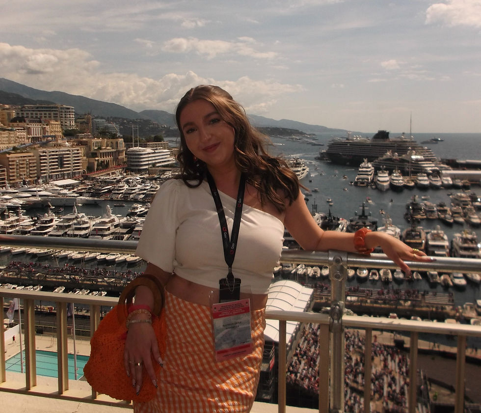 Woman smiling on balcony overlooking a marina with yachts. She wears a white top, orange checkered skirt, and carries an orange bag. Sunny day.