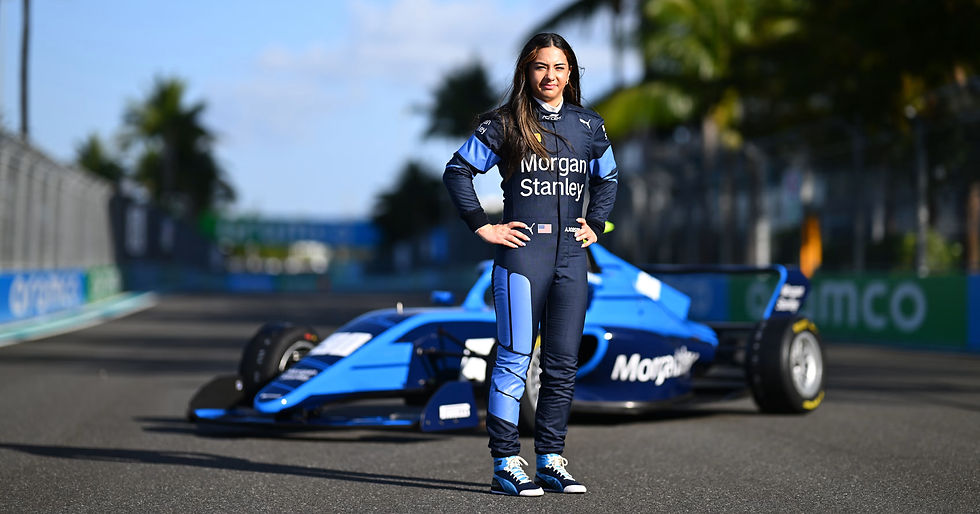 Woman in a blue Morgan Stanley racing suit stands on a track in front of a blue Formula car, hands on hips.