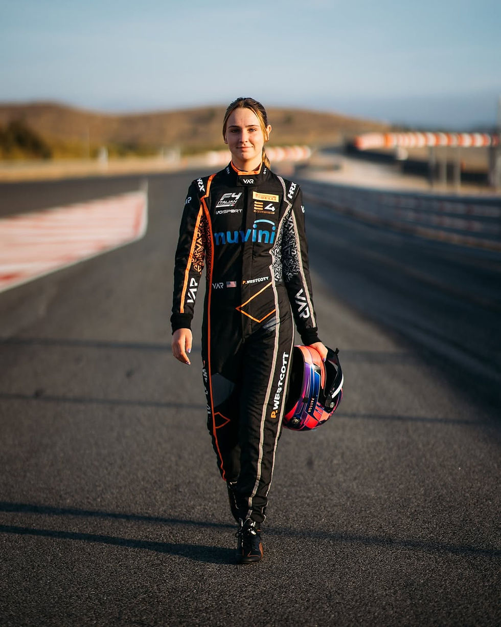 Race car driver in black suit walks on track holding helmet. Background shows blurred racetrack and landscape. Bright and determined mood.