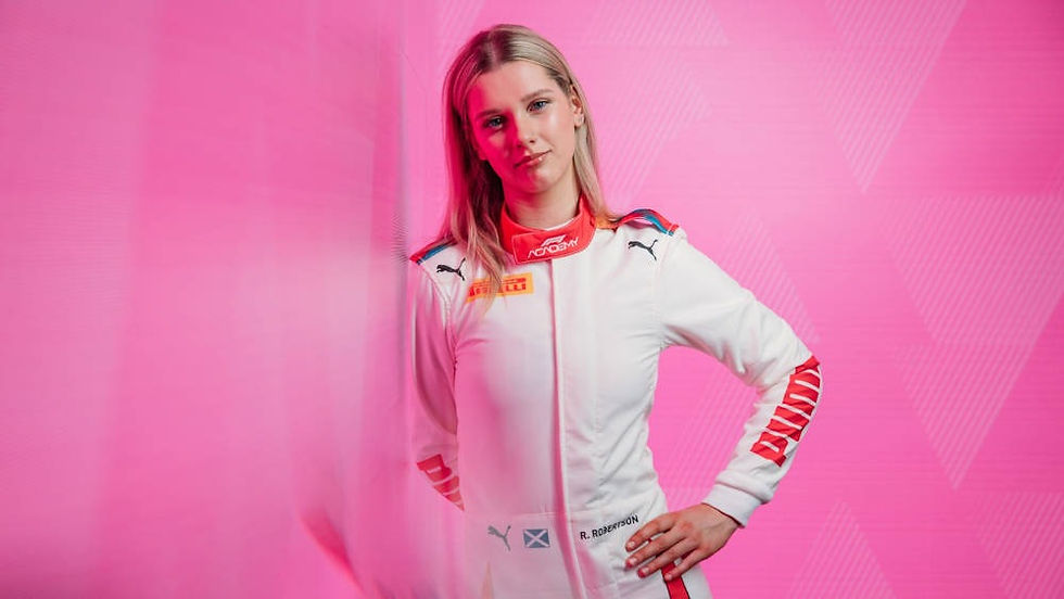 Person in a white racing suit with logos stands confidently before a pink background with subtle geometric patterns.