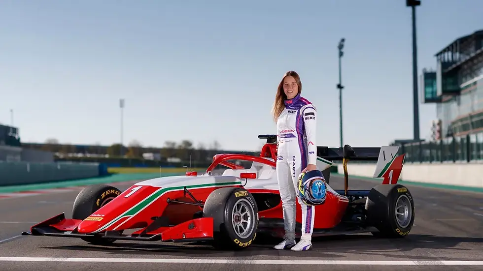 Female race car driver stands beside a red-and-white W Series car on a racetrack, holding her helmet and smiling.