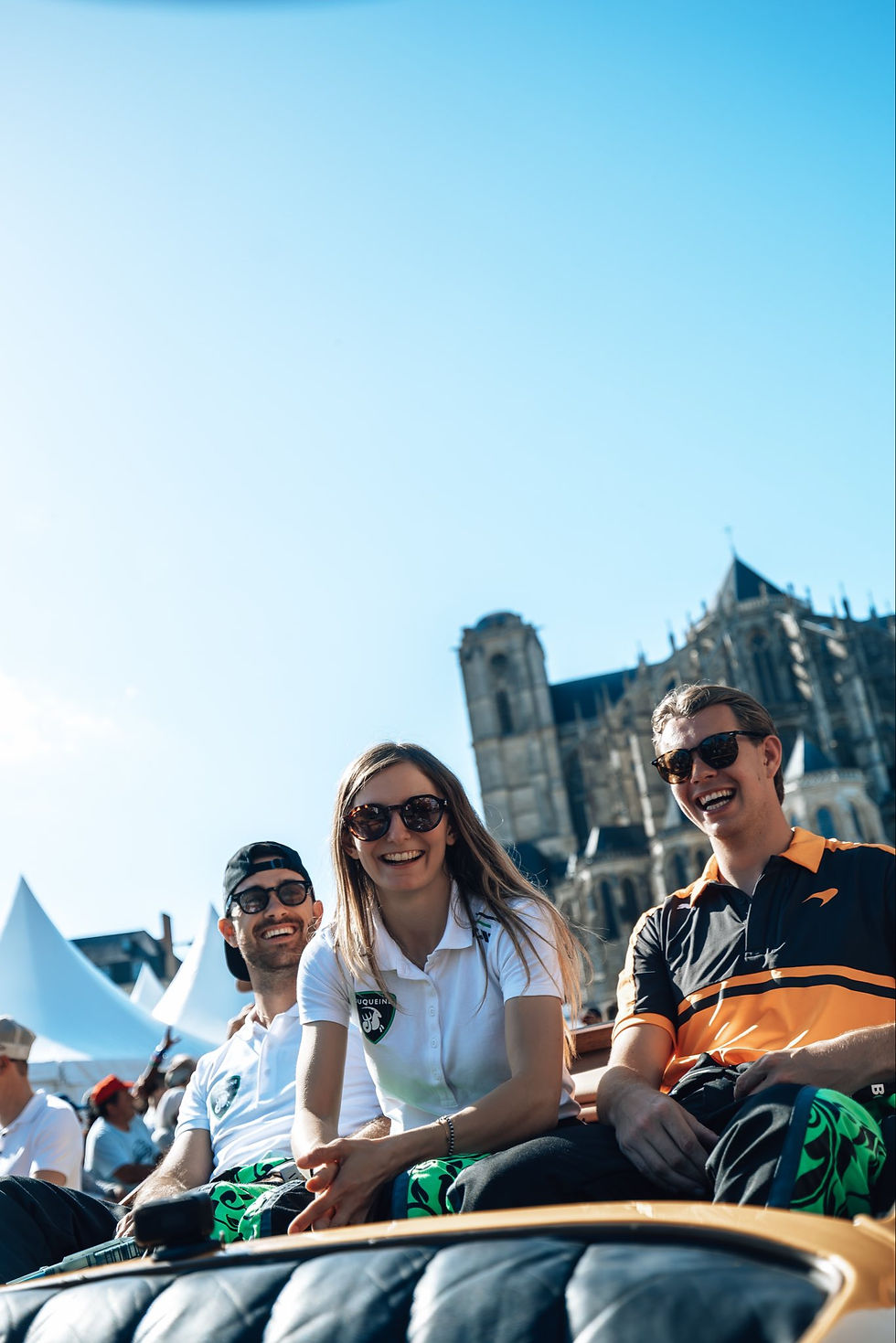 Three smiling young adults in sunglasses sit outdoors at a sunny event, with a cathedral and blue sky behind them.