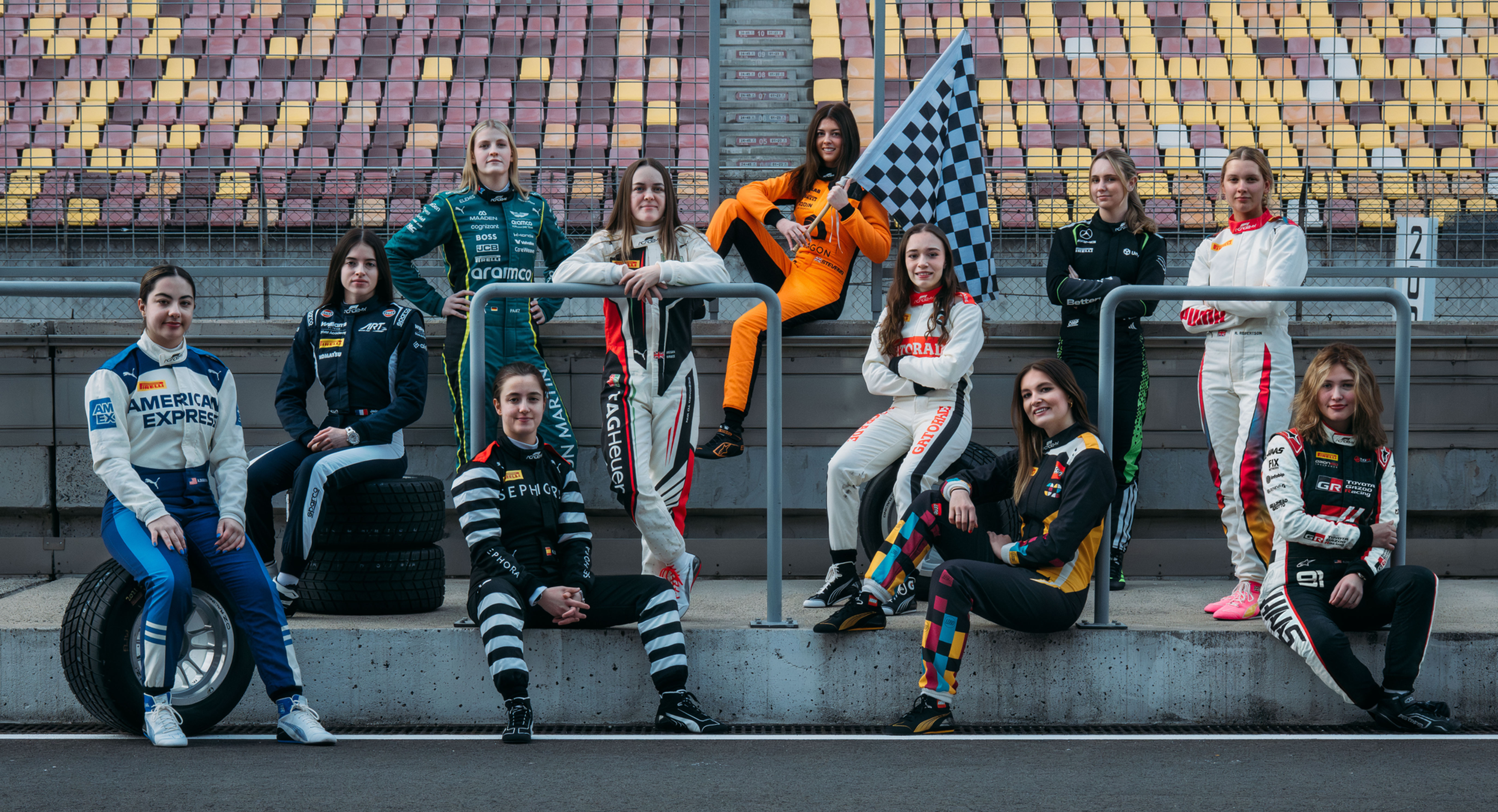 Group of women race car drivers in colorful suits pose on a racetrack, one holding a checkered flag, with empty stadium seats behind them