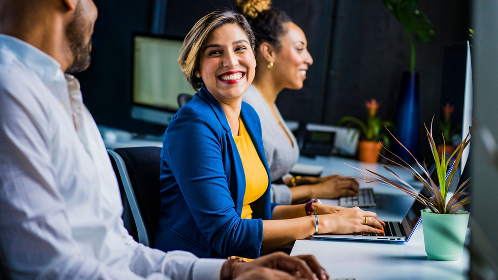 A close up picture of a woman in a yellow shirt with a blue blazer smiling at her coworker sitting next to her. They are working on their own laptop devices, showing independence and collaboration culture.