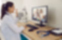 Online family doctor at desk with stethoscope smiling and waving hello at desktop computer