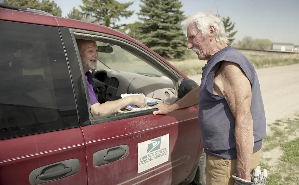 Rural letter carrier Roger McDonald, left, catches up with Edgar Clemens in rural Gering, Neb., on May 9, 2025 — McDonald’s first day back on the route after three months of medical leave. McDonald said he loves the sense of community fostered by the Postal Service, which he fears would disappear if his route was curtailed by cutbacks or eliminated by privatization. (Nick Loomis / The Midwest Newsroom)