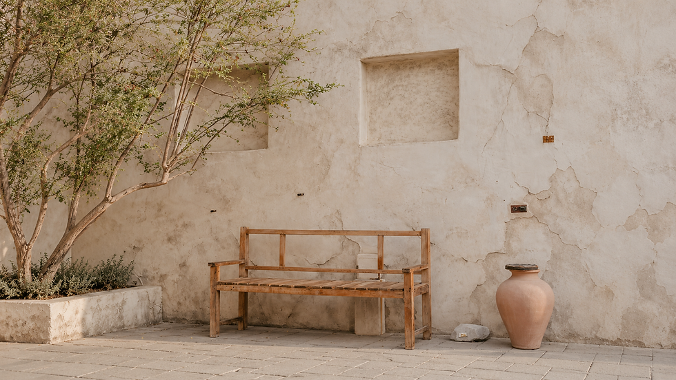 Weathered wooden bench, clay vessel, and leafy tree beside a textured plaster wall