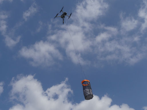 A drone carrying a cylindrical net with an orange rim flies against a blue sky with scattered clouds.