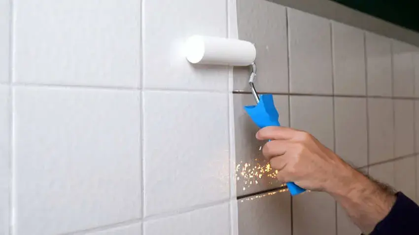Mano pintando azulejos blancos con rodillo y mango azul en baño. Ambiente limpio y ordenado, sin texto visible.