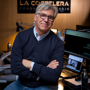 Un hombre de mediana edad, con gafas de pasta rectangulares y el pelo corto y canoso. va vestido con un jersey azul marino, una camisa blanca cuyo cuello sobresale por fuera del jersey y vaqueros. Está  sentado en un ambiente de oficina. Detrás de su cabeza, hay un cartel negro con letras troqueladas que ponen: La Coctelera Production Studio. 