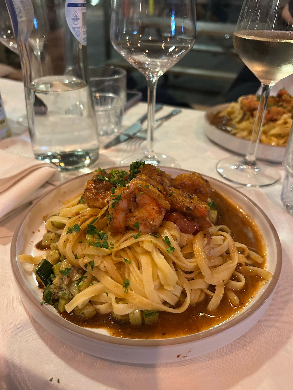 Plate of fresh tagliatelle coated in prawn bisque, topped with sautéed prawns, diced zucchini, and tomato concassé, garnished with chives and lemon zest.