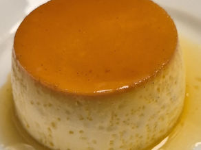 A golden crème caramel flipped onto a white plate, glossy caramel sauce pooling around the silky custard. A spoon rests nearby, and the soft reflection of candlelight hints at a dinner just shared around the table.