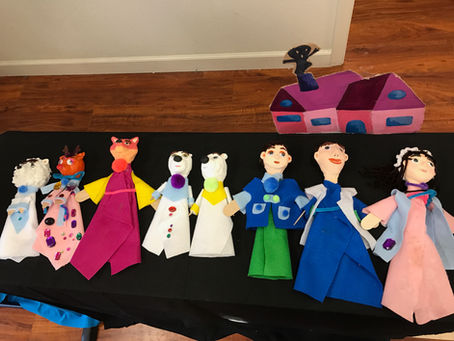 Colorful puppets in costumes on a black table, set against a wooden floor. A pink and purple house cutout is in the background.