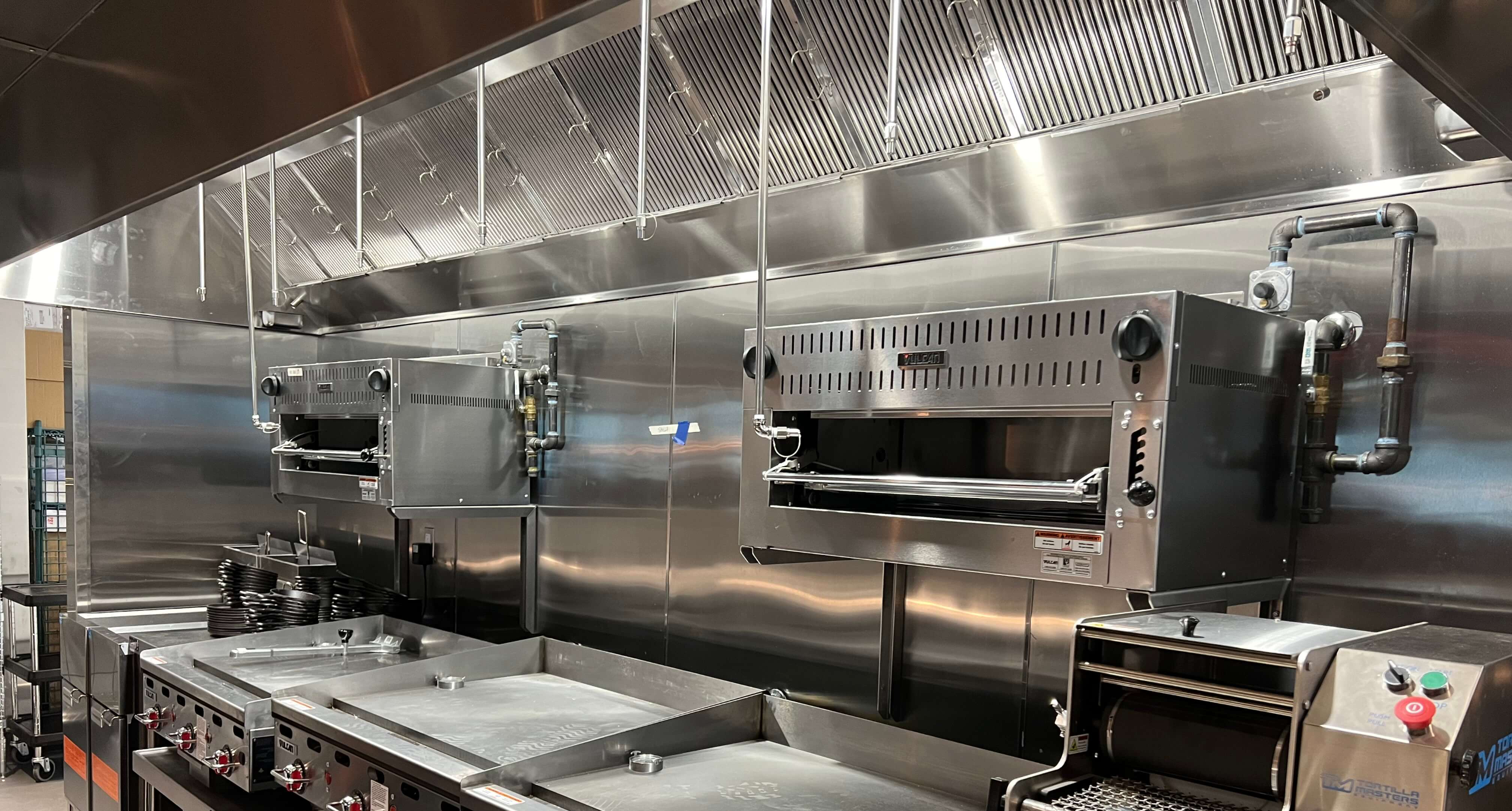 Industrial kitchen with stainless steel appliances, including grills, ovens, and ventilation hoods. Metal surfaces shine, creating a sleek look.