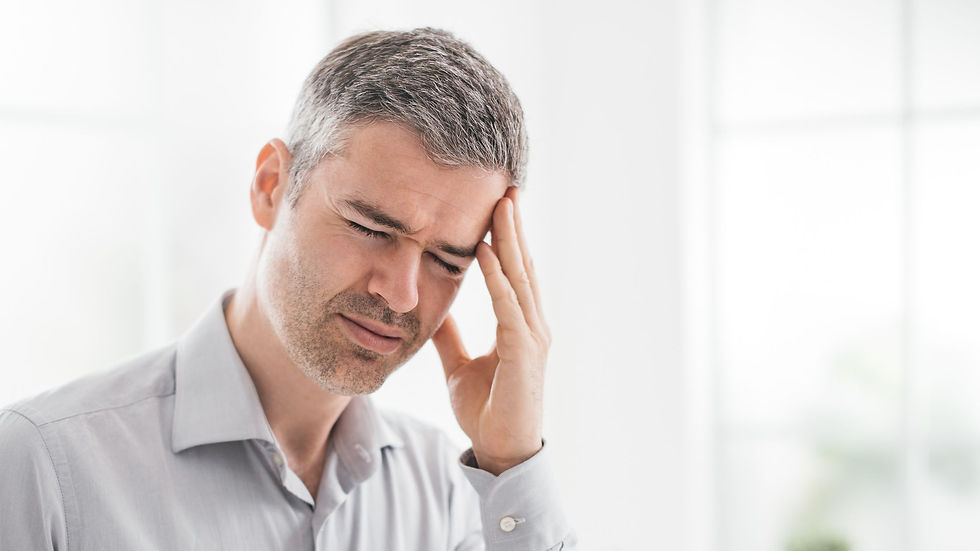 Man with gray hair holding head, eyes closed, appearing in pain. Wearing a gray shirt in a bright, blurred indoor setting.