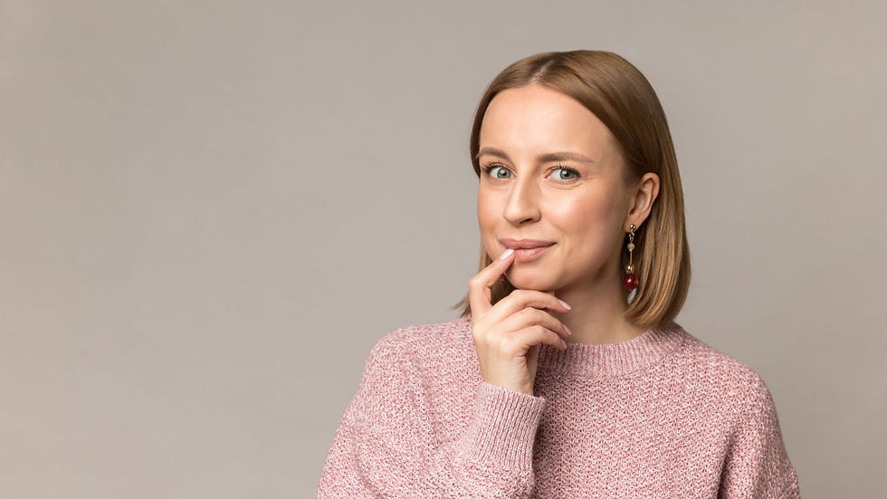 A woman in a pink sweater smiles subtly, touching her chin, against a plain beige background. She wears red earrings. The mood is thoughtful.