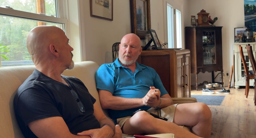 Two bald men sit on a couch talking in a sunlit living room with wood floors and furniture.