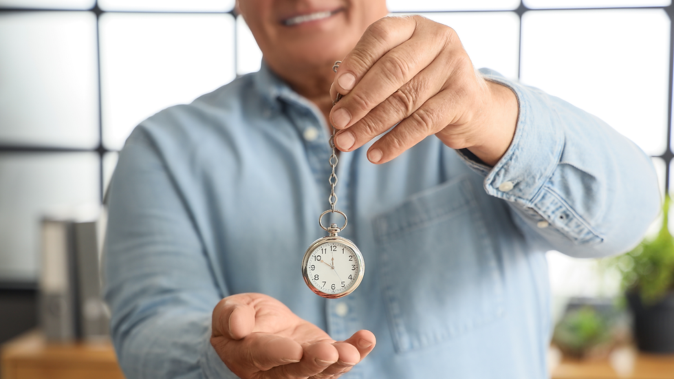 Man in a blue shirt holds a suspended pocket watch, smiling, against a bright indoor background. Focus on watch; calm atmosphere.