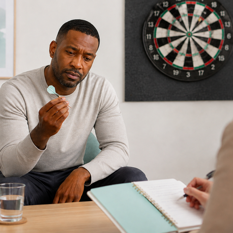 Man holds a dart while talking with a counselor in a calm office, with a dartboard on the wall and a notebook in the foreground.