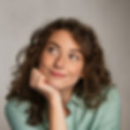 A young woman with curly brown hair, wearing a light green shirt, smiling thoughtfully with her chin resting on her hand. She has a relaxed and dreamy expression, looking slightly upwards.