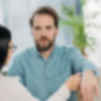 A man with a beard and a serious expression wearing a light blue shirt. He is undergoing a hypnosis session, with a woman's hand holding his arm, gently guiding it. A potted plant is visible in the background.