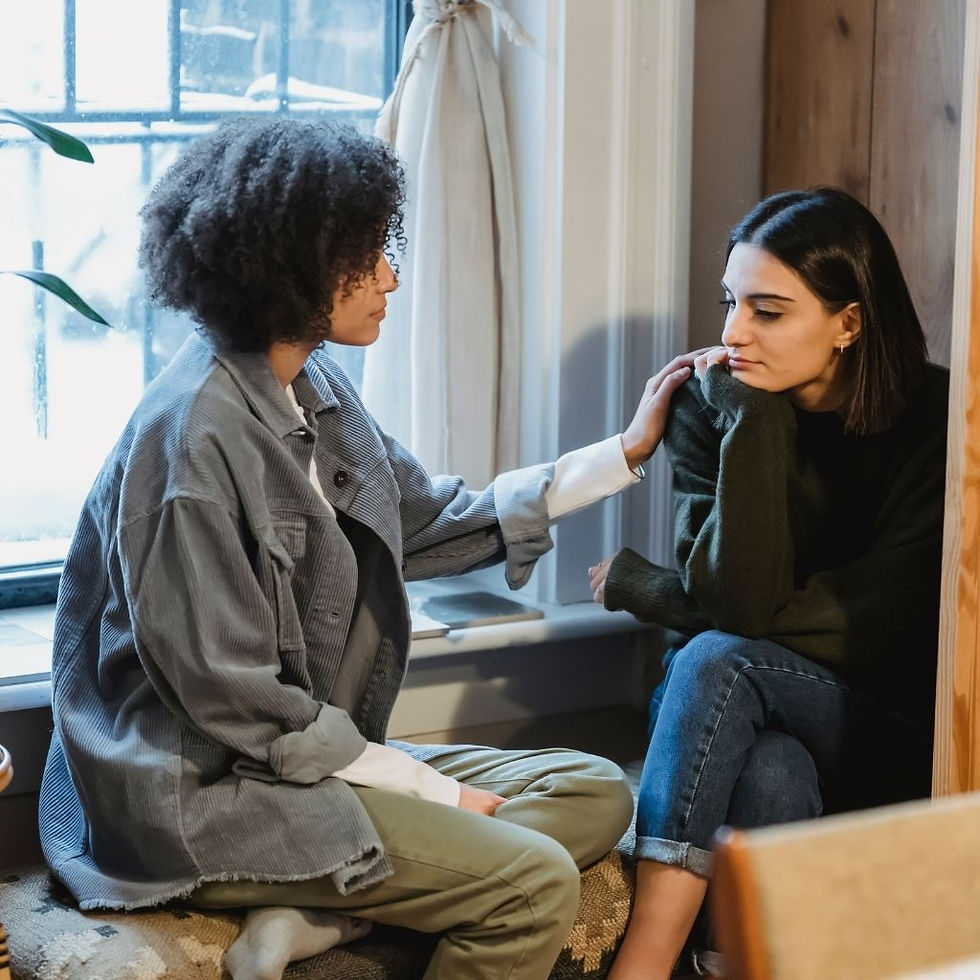 Two young women are sitting together indoors. One woman, with curly hair, is gently comforting the other woman, who has straight hair and looks sad. They are seated by a window, with natural light coming in.