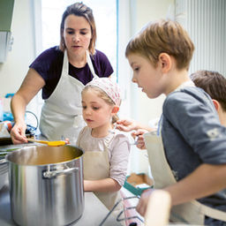 Eine Frau steht mit drei Kindern, alle mit Schürzen bekleidet, in einer Küche vor einer Kochzeile, vor ihnen ein großer Topf. Mit einem Löffel hebt sie Essen aus dem Topf, während die Kinder sie dabei beobachten.