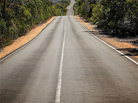 A long, straight two-lane road stretches toward the horizon through Australian bushland, with dense green shrubs and trees lining both sides. The road features a white center line and orange-red gravel shoulders. The asphalt undulates over multiple rolling hills creating a wave-like effect, with the road appearing to rise and fall dramatically in the distance before disappearing over the final crest against a pale blue sky.