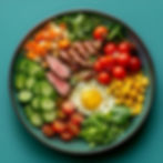 Colorful balanced meal bowl with sliced steak, sunny-side-up egg, cherry tomatoes, cucumbers, corn, leafy greens, diced sweet potato, and cheese on a green plate.