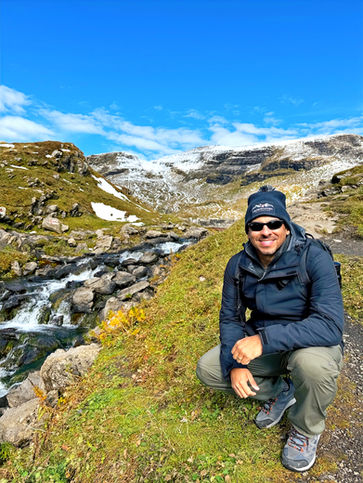 Man crouching on a path by a stream with snowy mountains