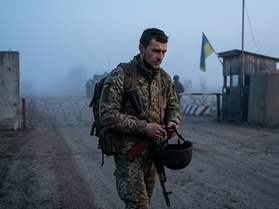 Український військовий у піксельному камуфляжі з рюкзаком та автоматом іде по ґрунтовій дорозі крізь туманний блокпост. Він тримає шолом у руках перед собою і дивиться вниз із втомленим виразом обличчя. На задньому плані видно бетонні блоки та український прапор на дерев'яній сторожці.