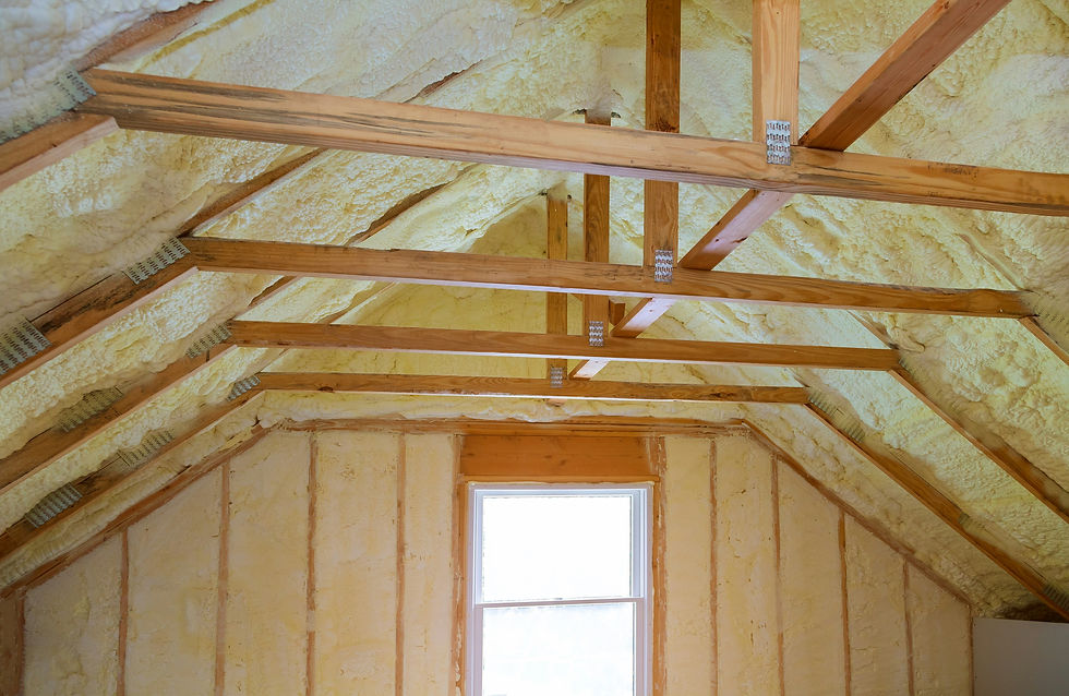 Wooden rafters and yellow foam insulation cover an attic ceiling. A window allows natural light, creating a warm, unfinished space.