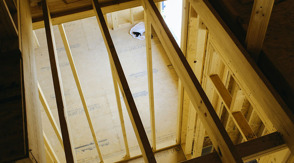 Wooden framing of a house under construction, viewed from above. Sunlight highlights beams and plywood floor. Text on plywood visible.