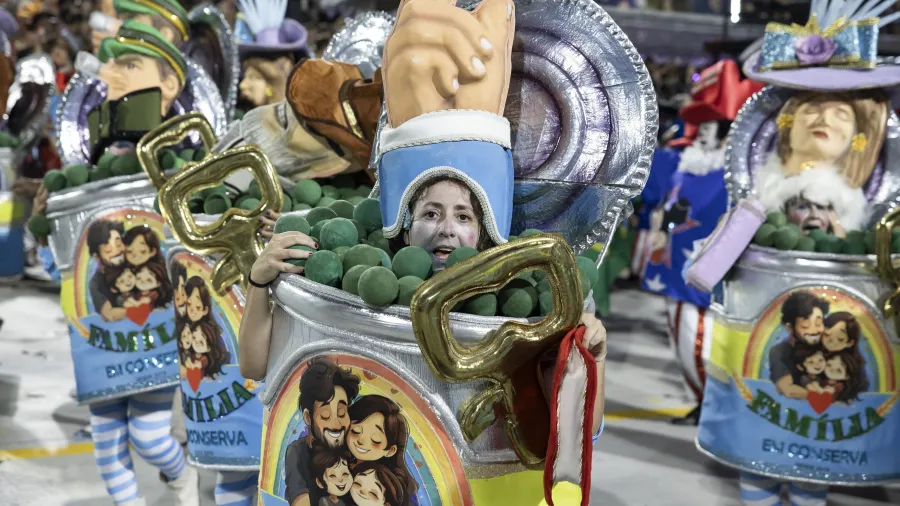 Desfilante em um carro alegórico de Carnaval fantasiado de lata de conserva, com tampa metálica aberta e bolas verdes dentro, levando ao peito a ilustração de uma família com a palavra “família”, enquanto outras “latas” semelhantes aparecem ao fundo na avenida.