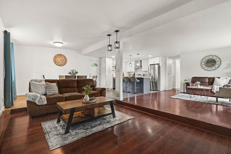 Modern living room with brown sofas, wooden tables, and white walls. A clock hangs on the wall, plants adorn tables, and a kitchen is visible.
