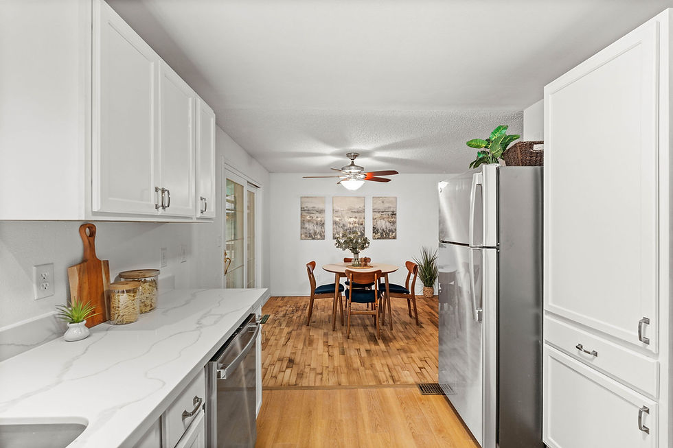 White kitchen with marble countertops, wooden cutting board, and glass jars. Dining area with round table, chairs, large plant, and artwork. Cozy vibe.