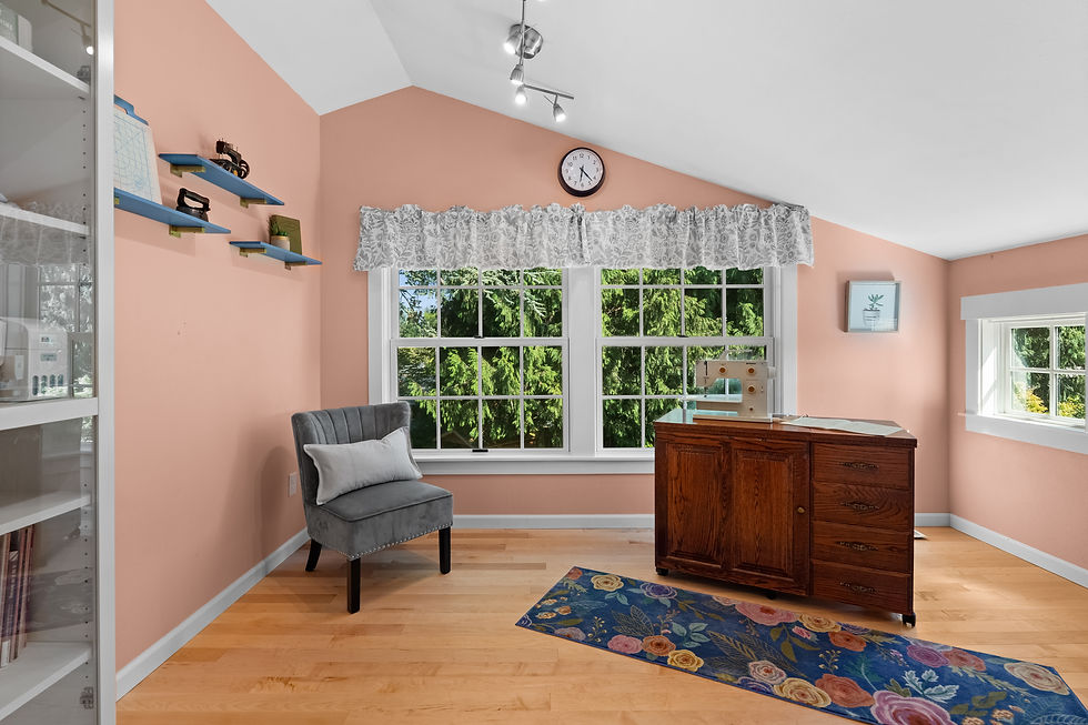 Bright sewing room with pink walls, gray chair, vintage cabinet, sewing machine, shelves, floral rug, and sunlit windows.