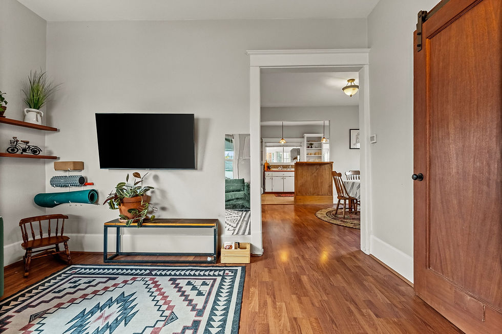 Cozy room with a patterned rug, mounted TV, plants, and shelves. Opens to a kitchen area with wooden floors and warm lighting.