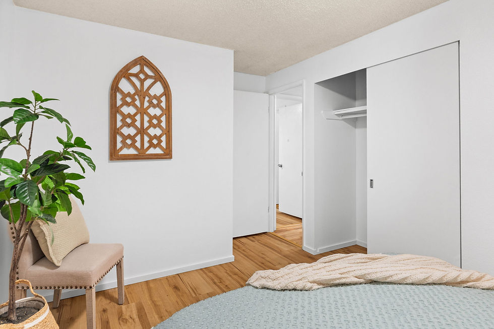 Simple bedroom with white walls and wooden floor. A bed with a blanket, a chair with a cushion, potted plant, and decorative wall panel.