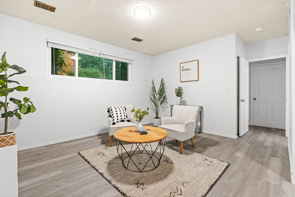 Cozy rec room with two beige chairs, round wooden table, plants, and wall art saying "home." Light wood flooring and window view.