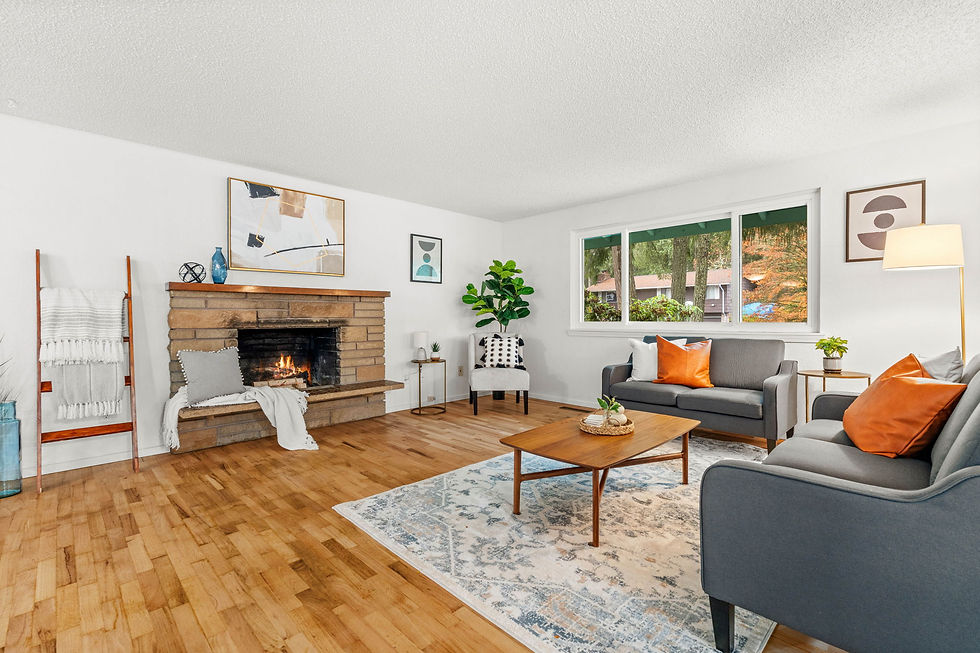 Cozy living room with a lit fireplace, grey sofas with orange pillows, and a wooden table on a patterned rug. Large windows show a leafy view.