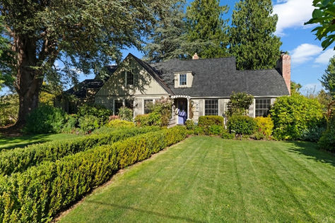 Sunny suburban house with a gray roof, lush hedges, and manicured lawn under tall trees and a blue sky